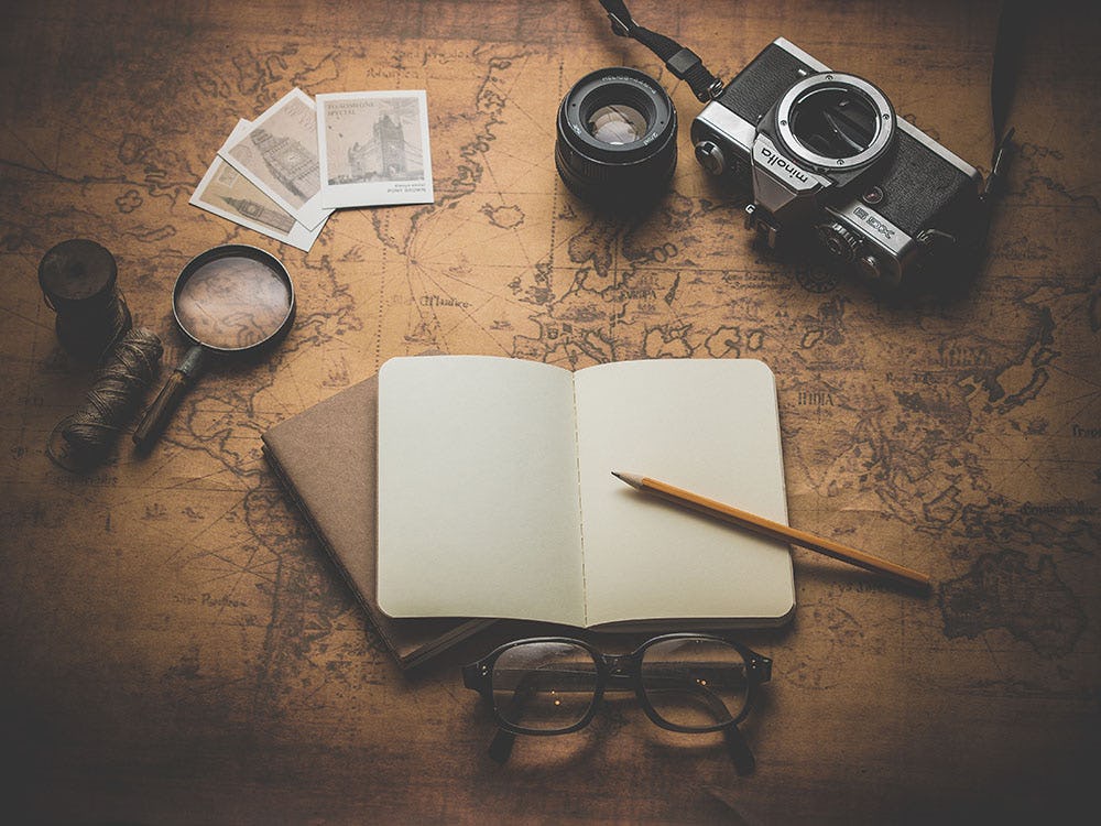 Un carnet de voyage ouvert et vide posé sur un vieux mappemonde marron. Entouré par une vieille loupe, de vieilles cartes postales, des lunettes et un appareil photo.